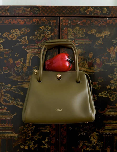 Olive green Loewe leather handbag with a red pepper inside, set against a dark ornamental wooden cabinet.