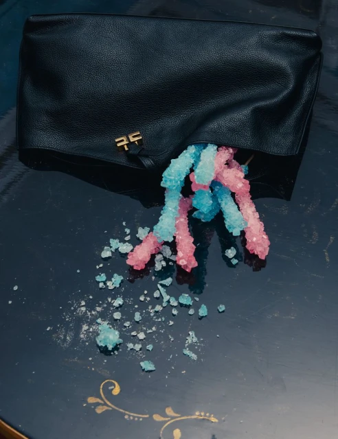 Black leather clutch bag with blue and pink rock candy spilling onto a dark surface with gold accents, overhead shot.