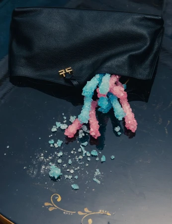 Black leather clutch bag with blue and pink rock candy spilling onto a dark surface with gold accents, overhead shot.