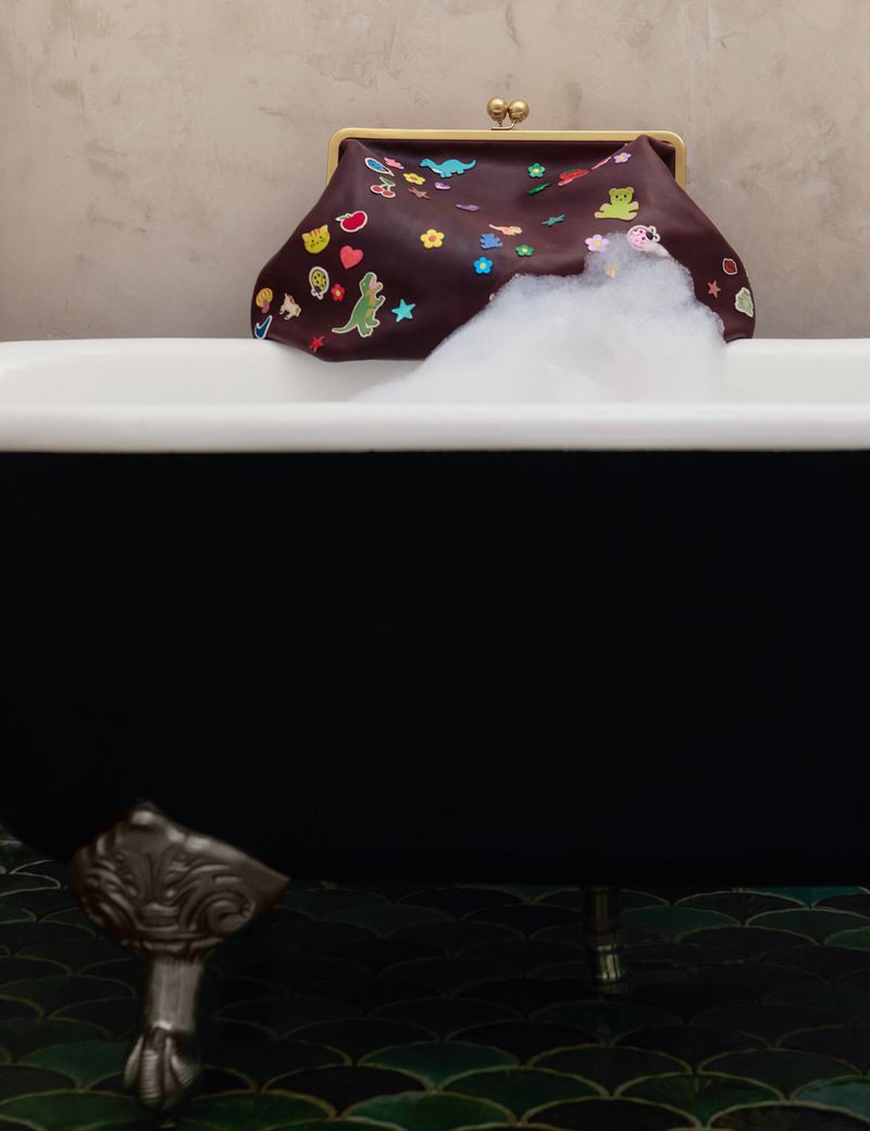 Close-up of a brown leather purse with colorful patches resting on a black bathtub filled with white foam.