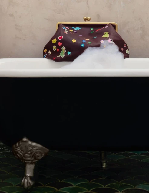 Close-up of a brown leather purse with colorful patches resting on a black bathtub filled with white foam.