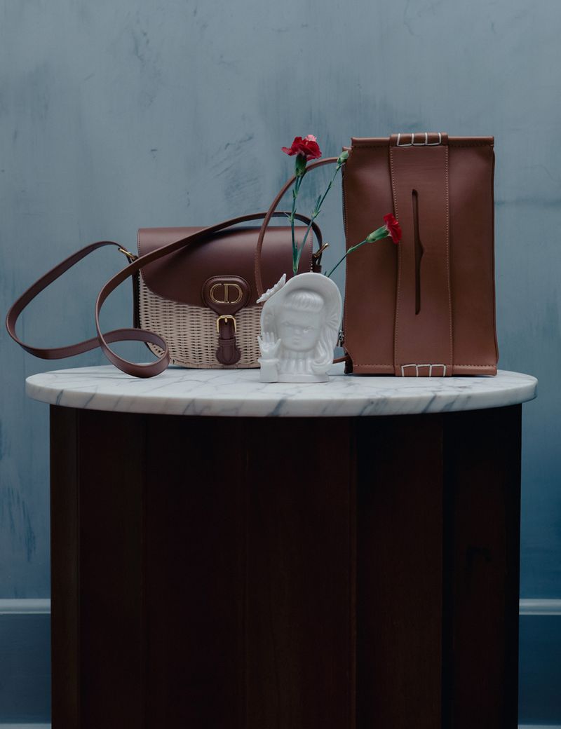 Still life of two leather handbags, a floral arrangement in a bust vase, on a round marble table against a textured grey-blue wall.