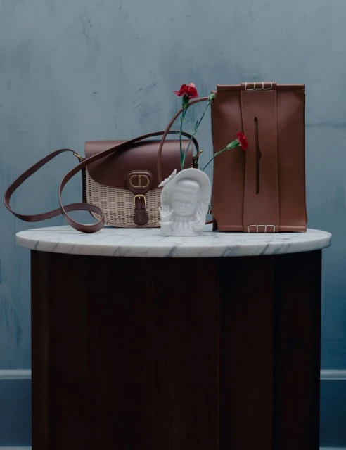 Still life of two leather handbags, a floral arrangement in a bust vase, on a round marble table against a textured grey-blue wall.