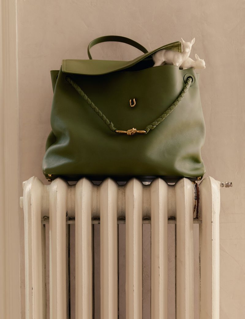 Olive-green leather purse sits atop a white radiator against a light beige wall. Two miniature figurines are tucked inside the purse.