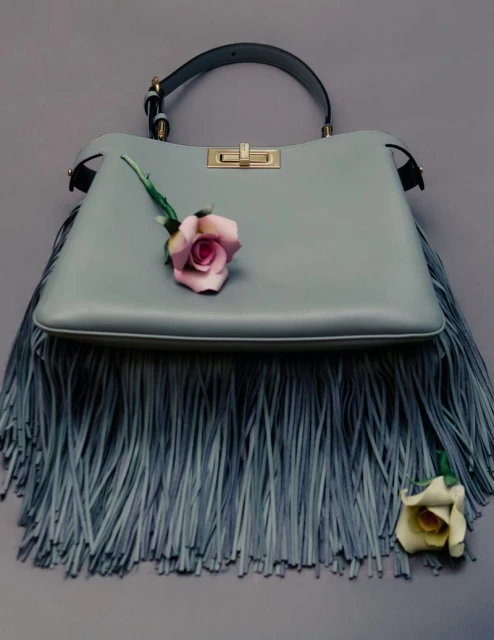 Light blue leather handbag with fringe detail and two roses, one pink, one white, against a gray background in a studio fashion shot.