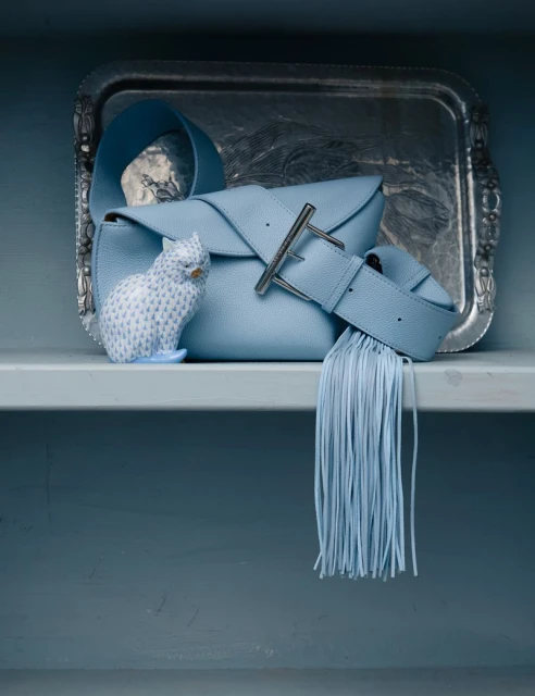 Light blue leather handbag with buckle detail, styled with a fringed strap, set on a silver tray with a patterned owl figurine.