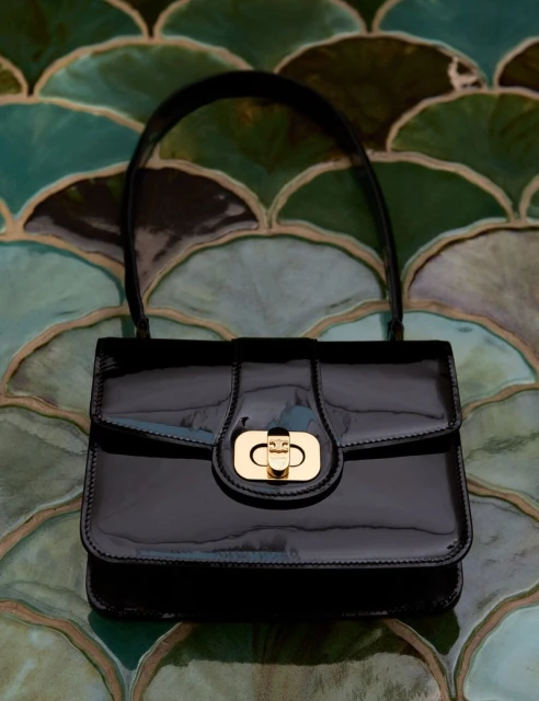 Close-up studio shot of a shiny black handbag with a gold clasp, set against a decorative patterned background of blue and green.