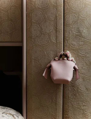 Pink textured leather handbag hanging on a patterned beige wall with embossed rose designs for Heroine Magazine Bag Bliss.