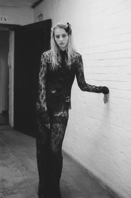 Full-length fashion shot of a woman in a black lace dress leaning against a white brick wall indoors under dim lighting.