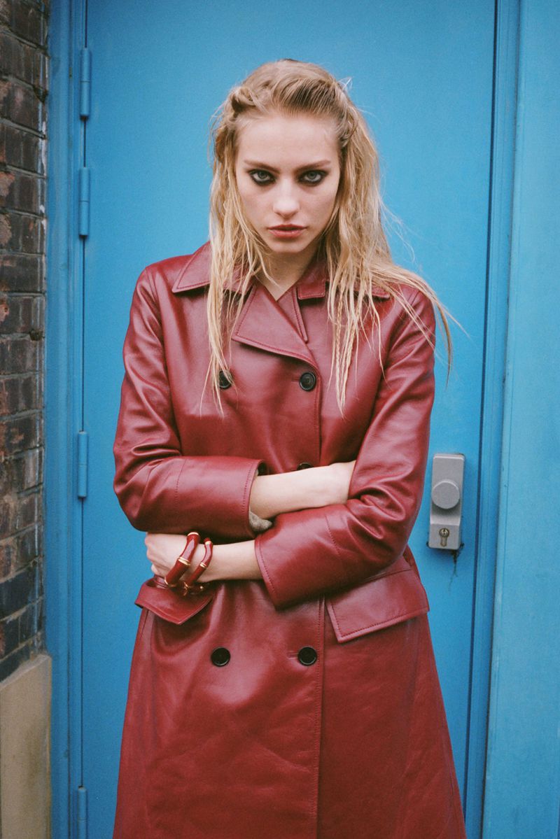A blonde woman with heavy makeup stands in front of a blue door wearing a red leather trench coat with a brick wall to the side.