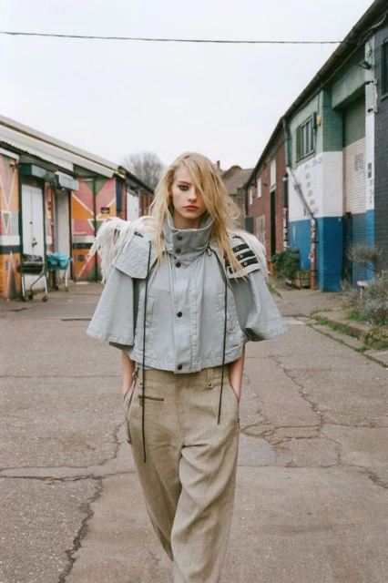 Fashion editorial shot of a blonde adult woman in a short-sleeved jacket and pants, walking on a cracked road between buildings.