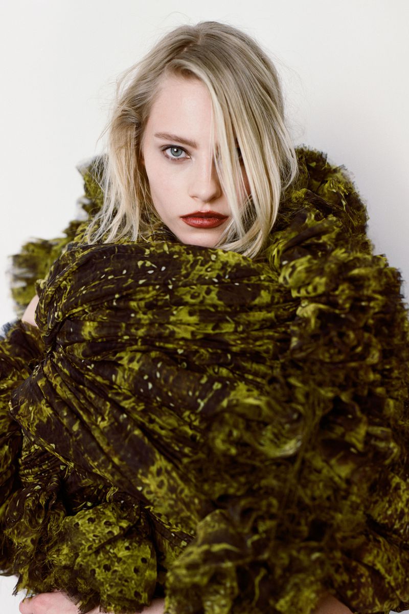Blonde woman wearing a green ruffled dress poses in a studio setting with soft lighting, showcasing fashion and beauty elements.