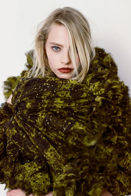 Blonde woman wearing a green ruffled dress poses in a studio setting with soft lighting, showcasing fashion and beauty elements.