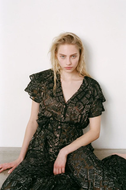 Young woman poses in a black, patterned jumpsuit against a simple white wall backdrop, fashion portrait, indoor studio setting, neutral expression.