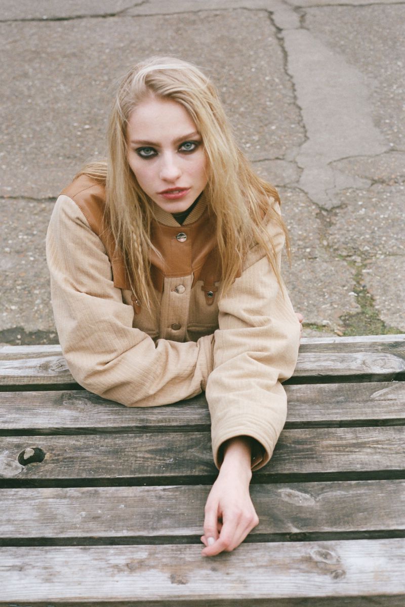 Young woman with long blonde hair and dramatic eye makeup, sitting on a weathered wooden bench outdoors, wearing a two-tone jacket.
