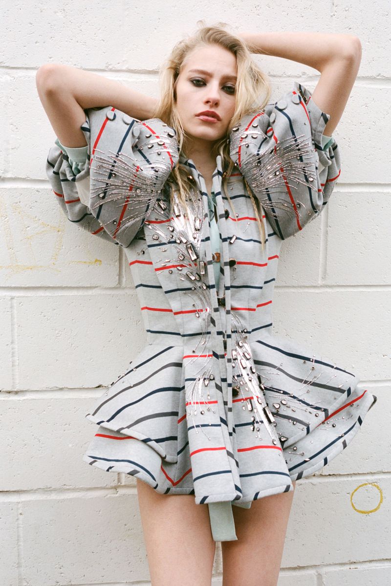 A female fashion model poses in a striped and embellished jacket in a studio setting, with a light-colored brick wall backdrop.