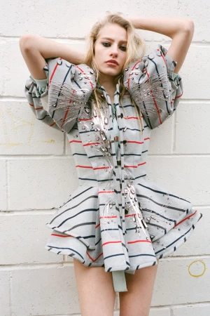 A female fashion model poses in a striped and embellished jacket in a studio setting, with a light-colored brick wall backdrop.
