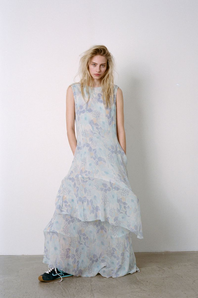 A solitary model poses in a light blue floral dress indoors against a white wall, accented by casual sneakers, for a fashion editorial.