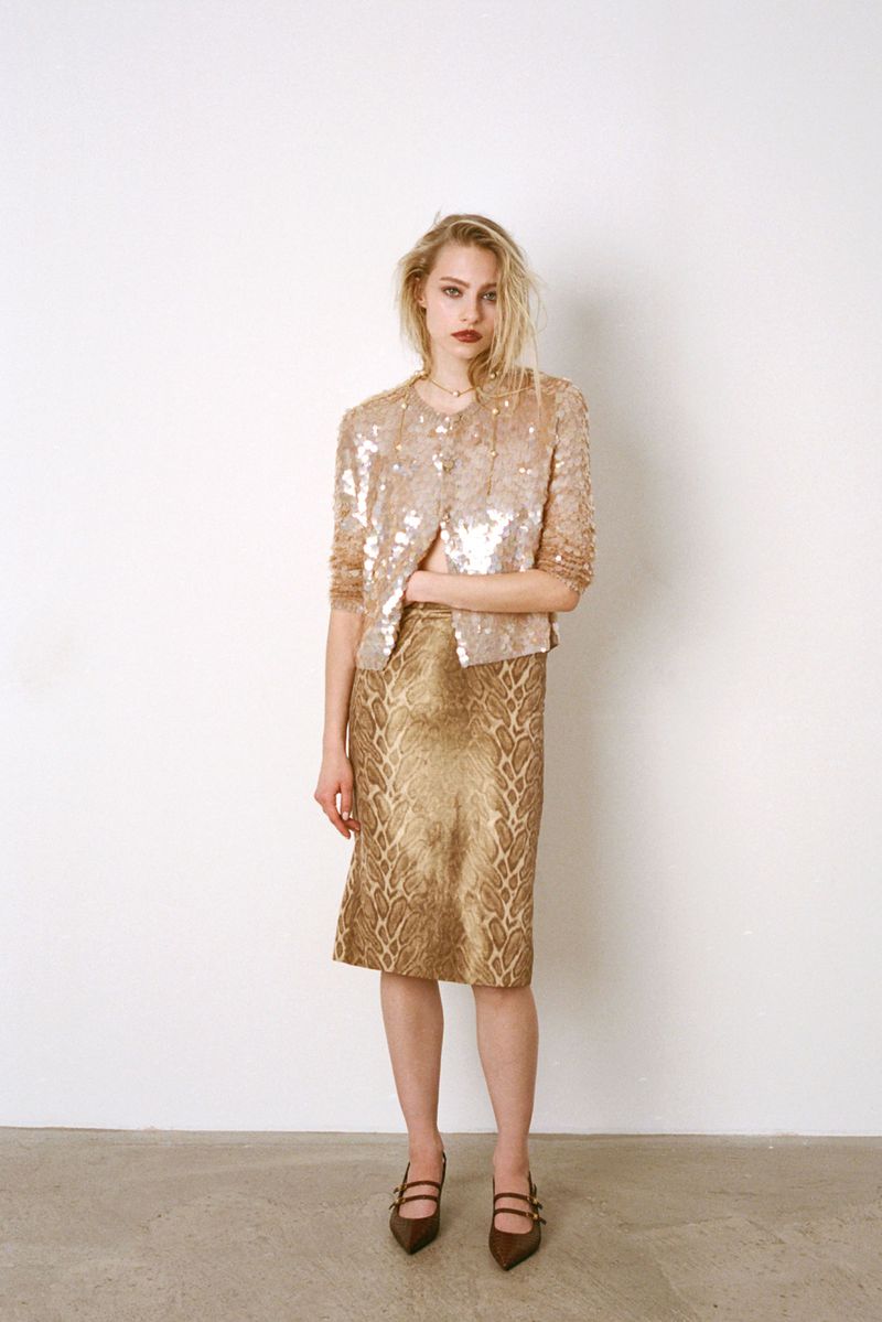 Full-length portrait of a woman in a shimmering, sequined jacket and patterned skirt, posing against a white wall in a fashion studio.