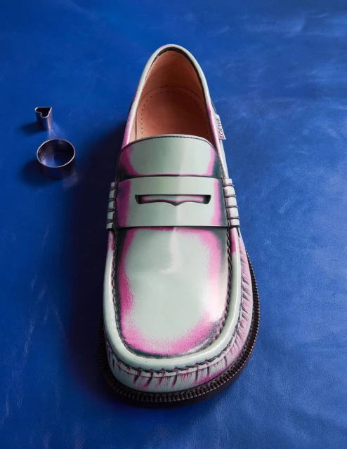Close-up of a glossy green and pink loafer with a thick sole on a textured blue surface with two metal cookie cutters nearby.
