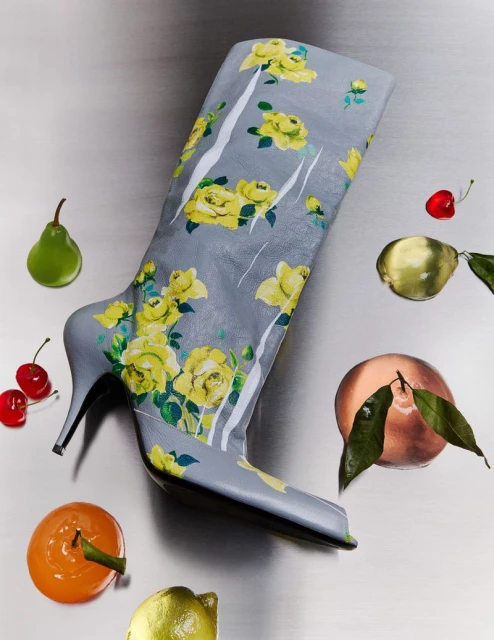 Grey knee-high boot with yellow floral design surrounded by pears, oranges, lemons, and cherries against a metallic surface.
