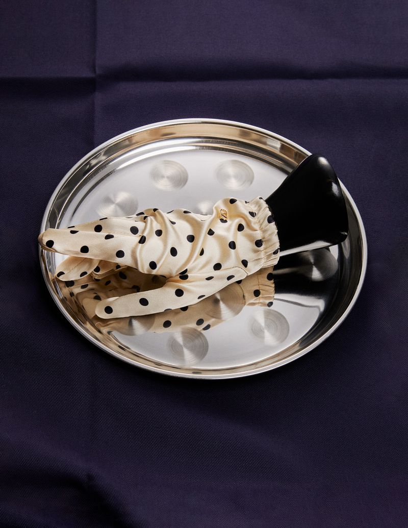 Elegant black heeled shoe and polka dot glove arranged on a reflective silver tray atop a dark blue fabric backdrop. 