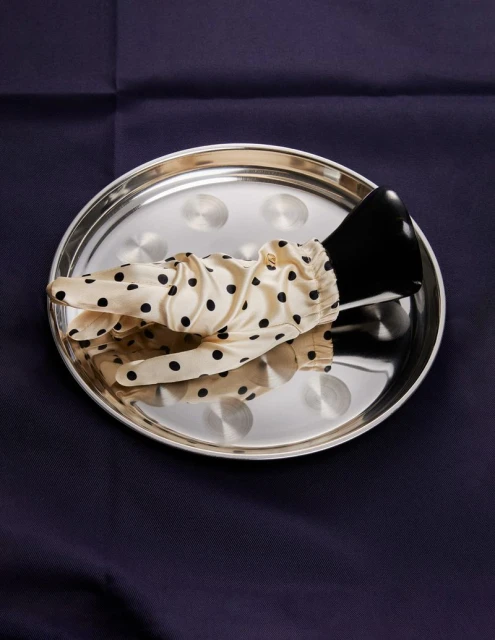 Elegant black heeled shoe and polka dot glove arranged on a reflective silver tray atop a dark blue fabric backdrop. 