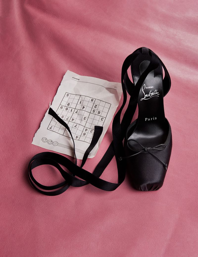 Black Christian Louboutin ballet heel with ankle ribbons next to a sudoku puzzle on pink textured surface in a studio.