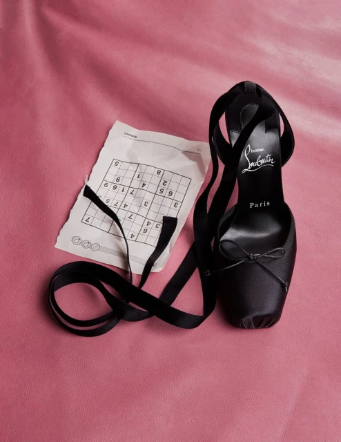 Black Christian Louboutin ballet heel with ankle ribbons next to a sudoku puzzle on pink textured surface in a studio.