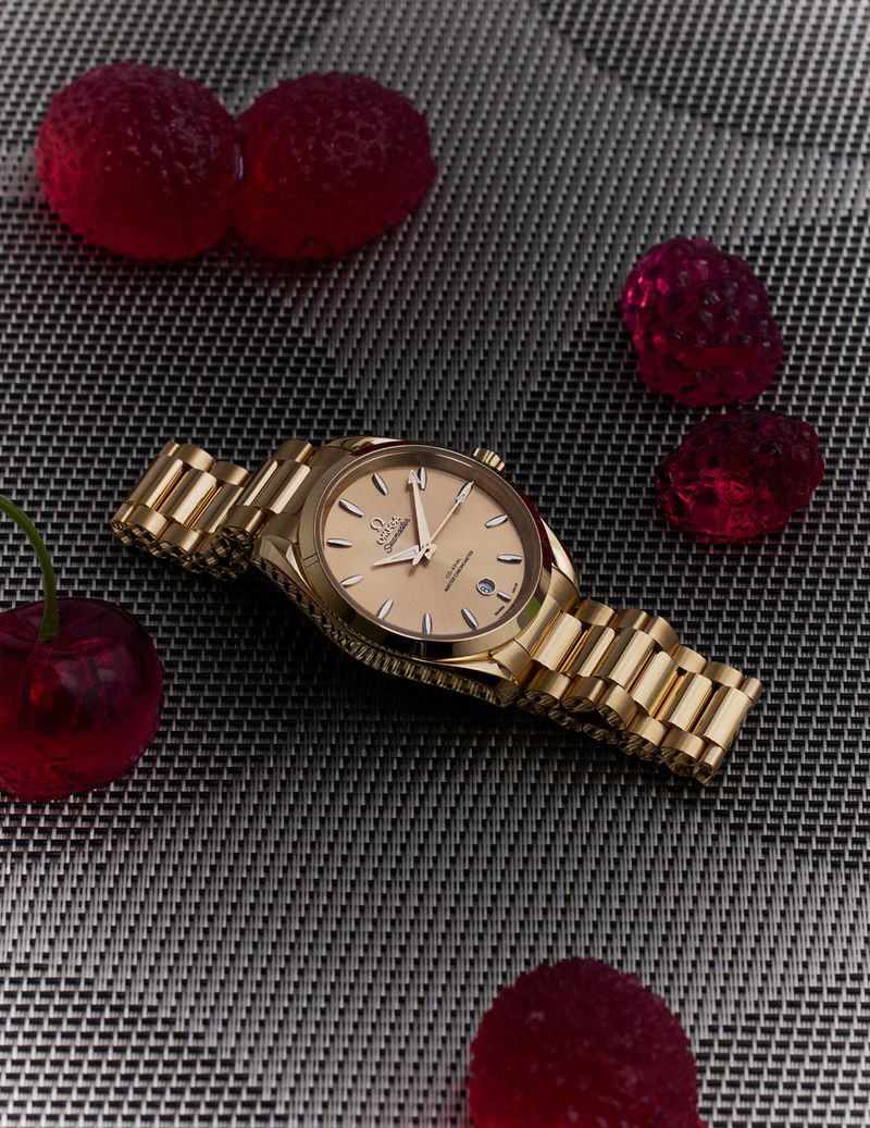Gold wristwatch with matching bracelet on a textured surface, surrounded by red gummy candy resembling berries and cherries.