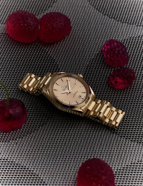 Gold wristwatch with matching bracelet on a textured surface, surrounded by red gummy candy resembling berries and cherries.