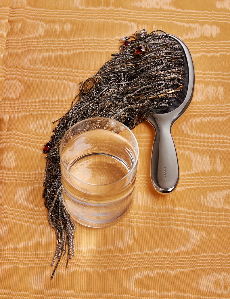 Still life shot featuring a clear glass of water, a silver hairbrush adorned with jewelry, resting on a textured orange fabric.