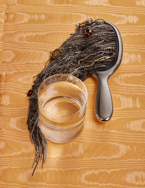 Still life shot featuring a clear glass of water, a silver hairbrush adorned with jewelry, resting on a textured orange fabric.