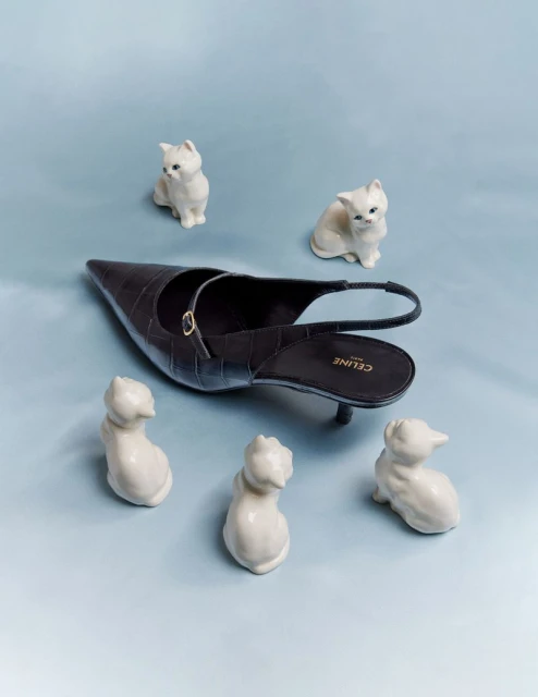 Dark brown slingback heel by Celine surrounded by five white ceramic cat figurines on a light blue backdrop in a studio fashion shot.