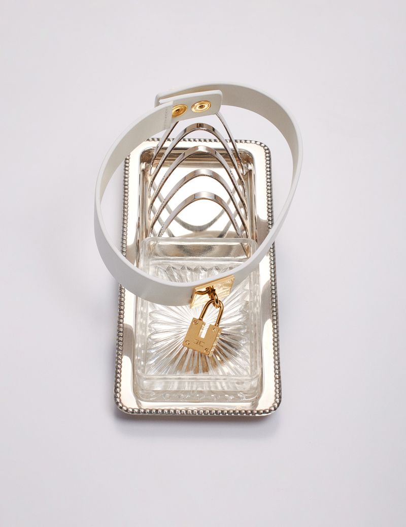 Silver plated toast rack with a white leather dog collar with gold padlock on a white background in a studio setting.