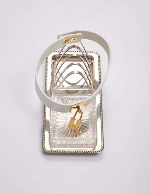 Silver plated toast rack with a white leather dog collar with gold padlock on a white background in a studio setting.