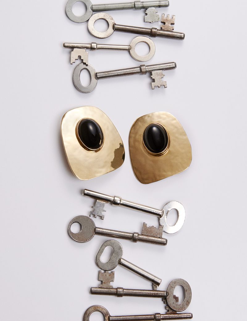 Overhead shot displays an arrangement of old metal keys and gold earrings with black stones on a clean white surface, creating a striking contrast.