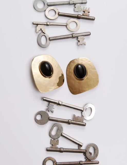 Overhead shot displays an arrangement of old metal keys and gold earrings with black stones on a clean white surface, creating a striking contrast.