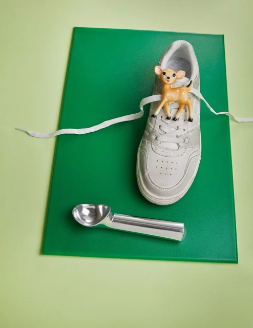 Still life composition: A white sneaker with a toy deer on top, an ice cream scoop, and a green surface on a light green background.