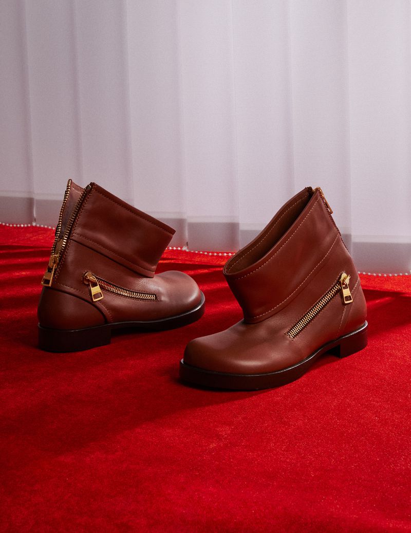 Two brown leather ankle boots with zipper details on a red carpet in a photo studio with soft window light creating a fashion product shot.