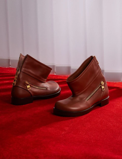 Two brown leather ankle boots with zipper details on a red carpet in a photo studio with soft window light creating a fashion product shot.