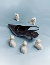 Celine black slingback kitten heel surrounded by five small white ceramic cat figurines on a pale blue surface