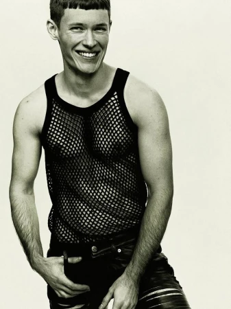 Close-up portrait of a male model wearing a black fishnet tank top and dark trousers against a plain light background.