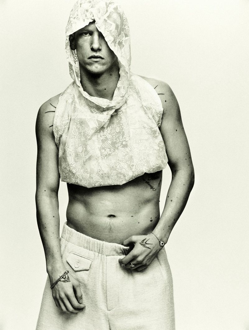 Monochrome portrait of a shirtless male model wearing white pants and a lace hoodie, showcasing fashion and style against a light background.