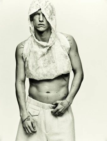 Monochrome portrait of a shirtless male model wearing white pants and a lace hoodie, showcasing fashion and style against a light background.