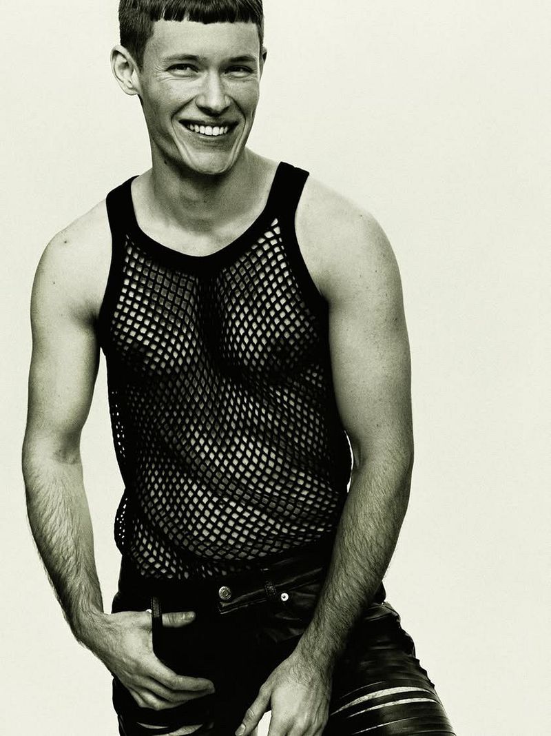 Close-up studio shot of a smiling adult male with short hair wearing a black fishnet tank top and leather pants against a plain background.