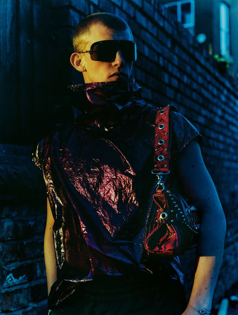 Stylish man with sunglasses and edgy clothing poses against a brick wall at night, accented by blue and yellow lighting.