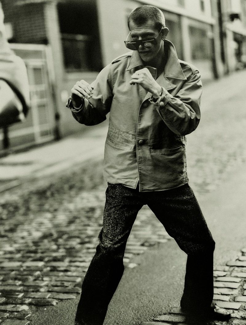 Monochrome shot of a man in sunglasses and a jacket in a defensive boxing pose on a cobblestone street, with buildings in the background.