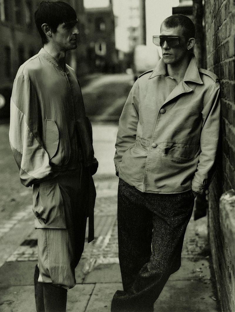 Two male models standing in an alleyway, one leaning against a brick wall wearing futuristic sunglasses and a jacket.