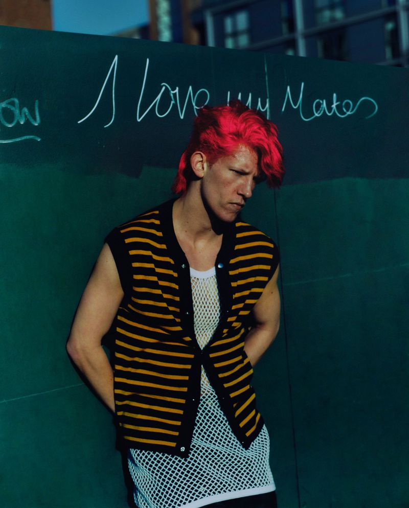 Man with red hair and striped vest leans against a dark green wall with white graffiti in an outdoor urban setting on a sunny day.